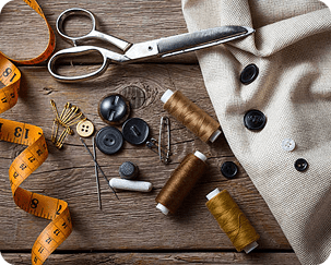 A sewing kit with scissors, measuring tape, needles, thread spools, buttons, pins, and fabric laid out on a wooden surface.
