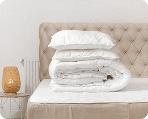 A bed with a tufted headboard holds a stack of two white pillows and a folded white comforter; a wicker lamp sits on a small round table nearby.