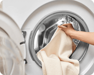 A person’s hand places a beige knitted sweater into the open drum of a front-loading washing machine.