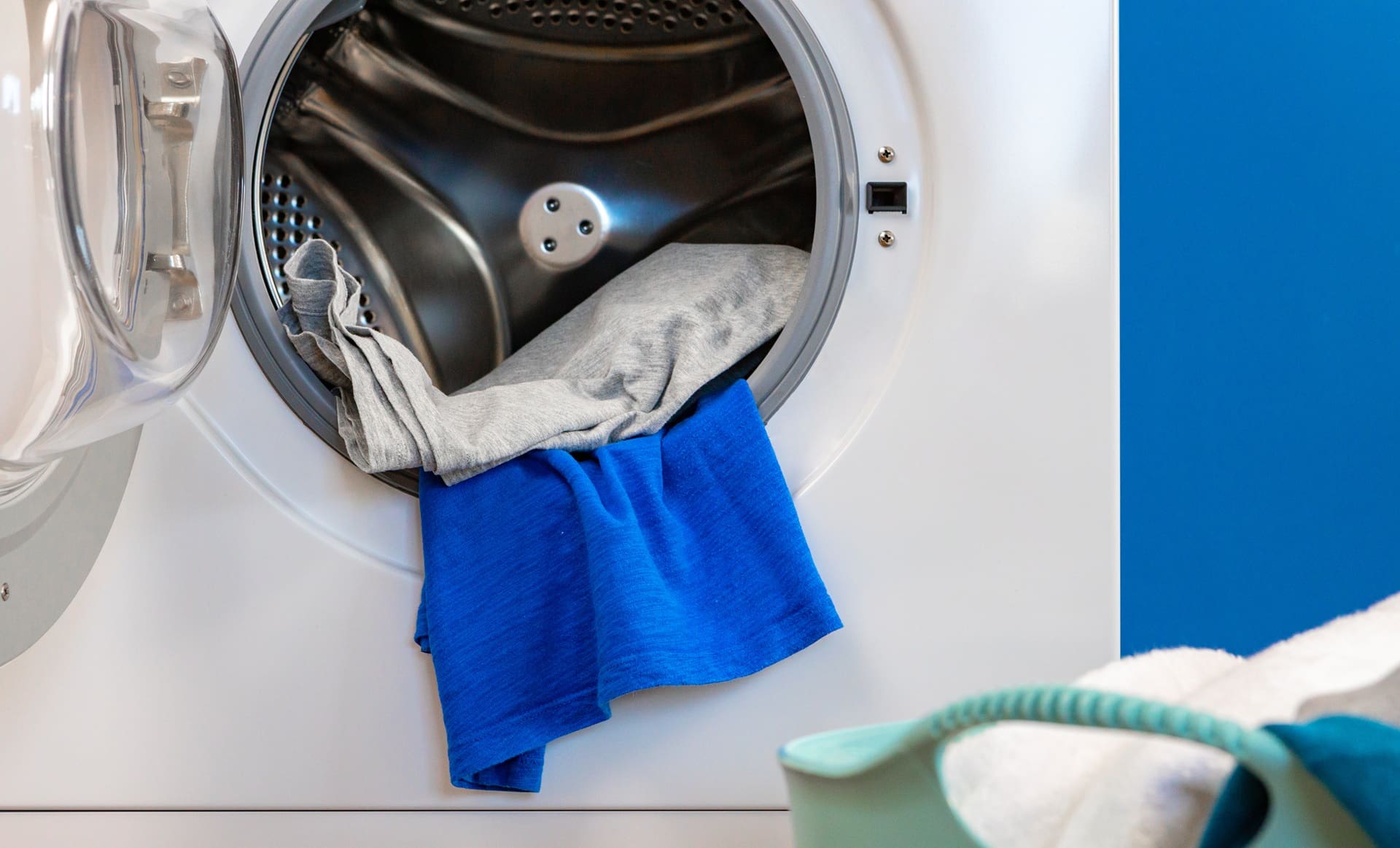 Front-loading washing machine with its door open, showing a grey and a blue garment hanging out. A laundry basket with towels is visible in the foreground.