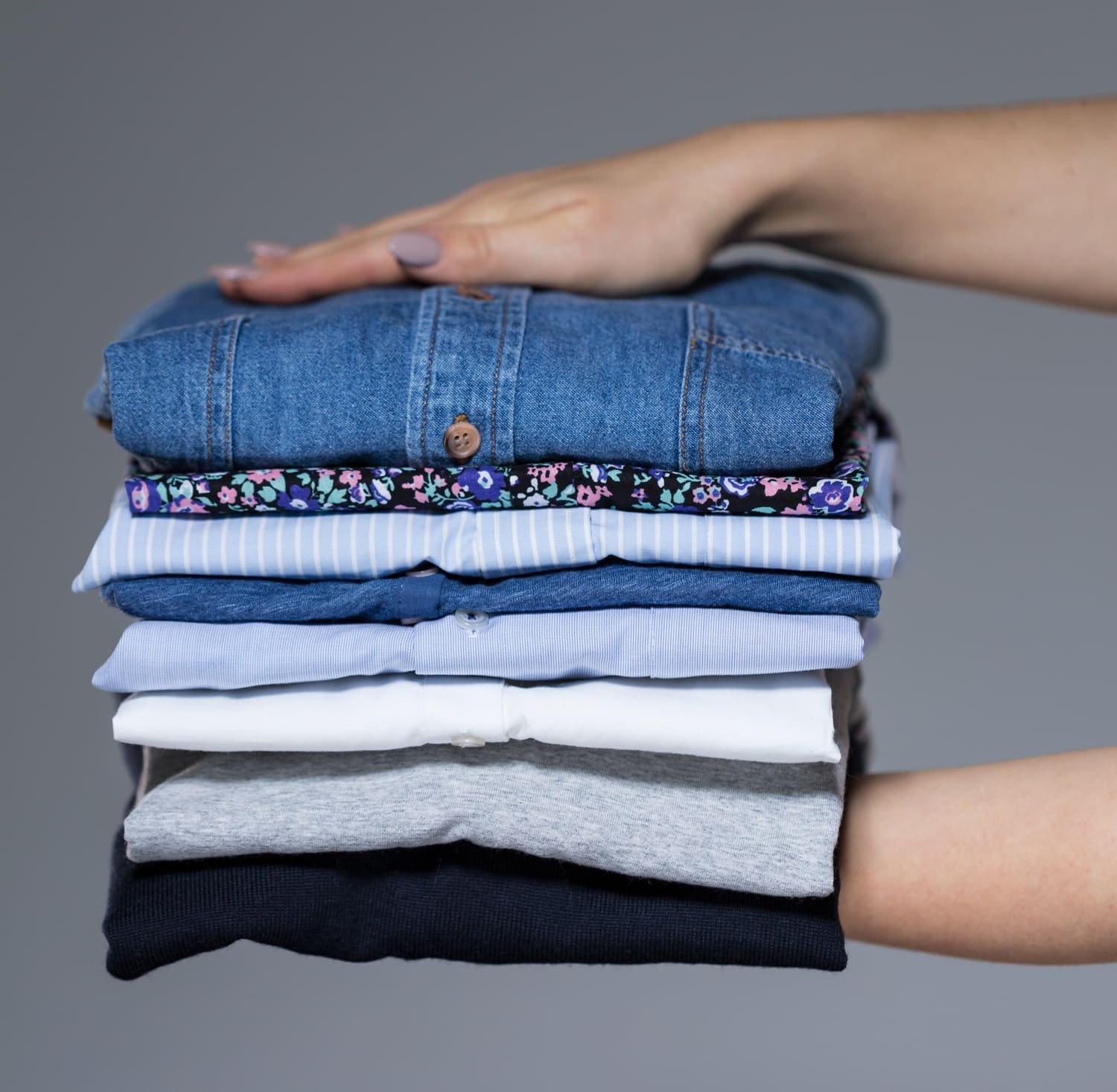 A hand holding a neatly folded stack of assorted shirts and tops in different colors and patterns against a plain background.