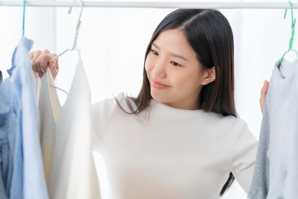 A woman stands in front of a clothing rack, holding and examining garments on hangers.
