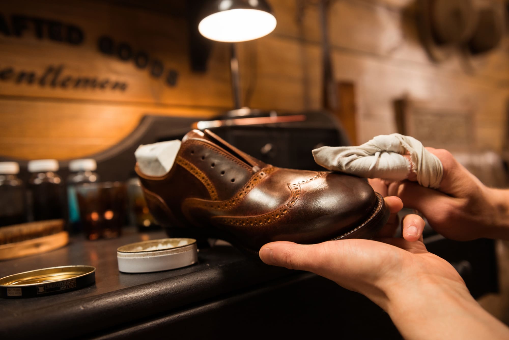A person polishes a brown leather dress shoe by hand, using a cloth and shoe polish, in a workshop setting under warm lighting.