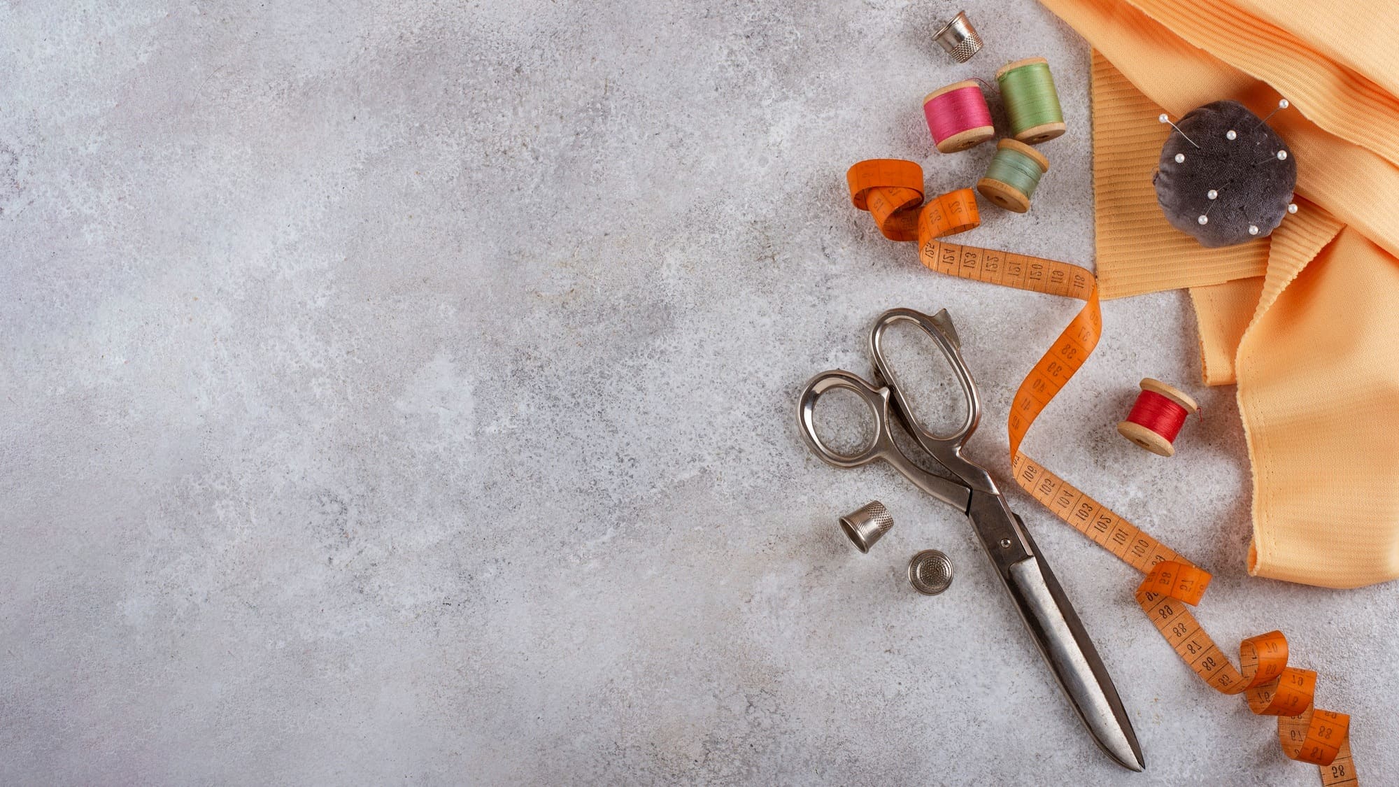 Scissors, thimble, measuring tape, spools of thread, pins, and folded orange fabric arranged on a gray textured surface.