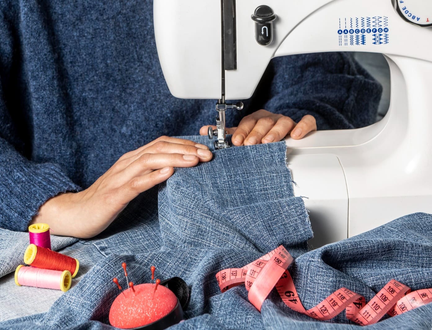 Person sewing blue fabric with a white sewing machine, surrounded by spools of thread, a pincushion, and a measuring tape.