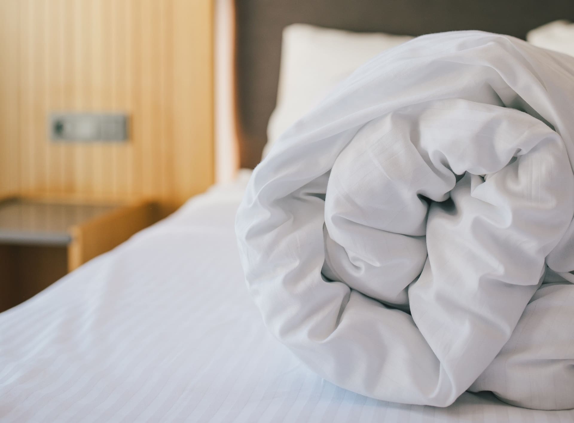 A close-up of a neatly rolled white comforter placed on a made bed with white sheets in a bedroom.