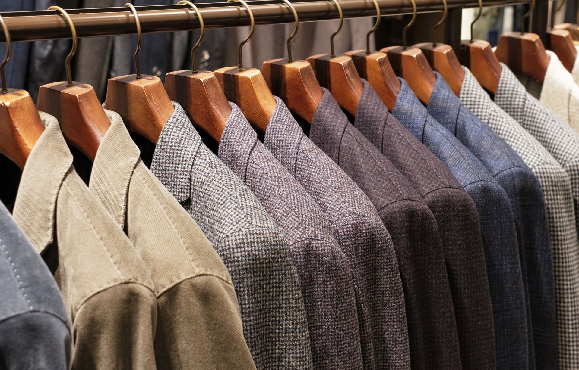 A row of men's suit jackets in various colors and patterns hanging on wooden hangers on a clothing rack.