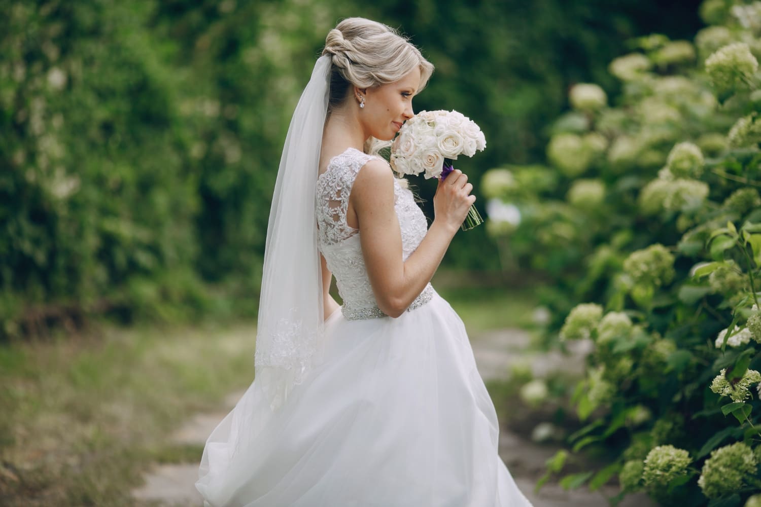 A bride in a white wedding dress and veil stands outdoors, holding a bouquet of white roses near green bushes.