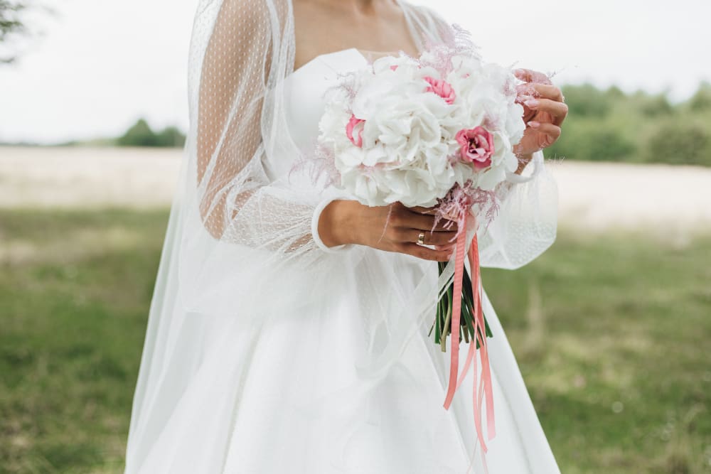 Bride in a white wedding dress holding a bouquet of white and pink flowers with ribbons, standing outdoors in a green field.