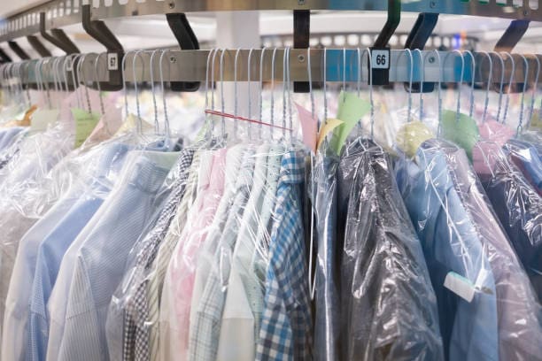 A row of freshly cleaned shirts and jackets covered in plastic hangs on a metal rack at a dry cleaning facility.