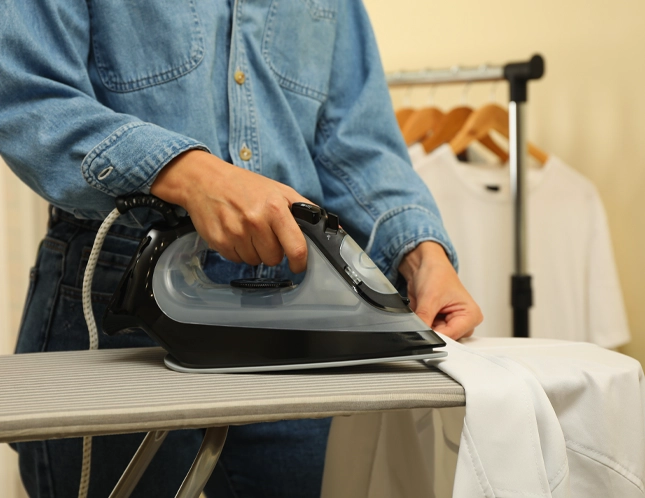 A person in a denim shirt irons a white garment on an ironing board. In the background, shirts hang on a rack.