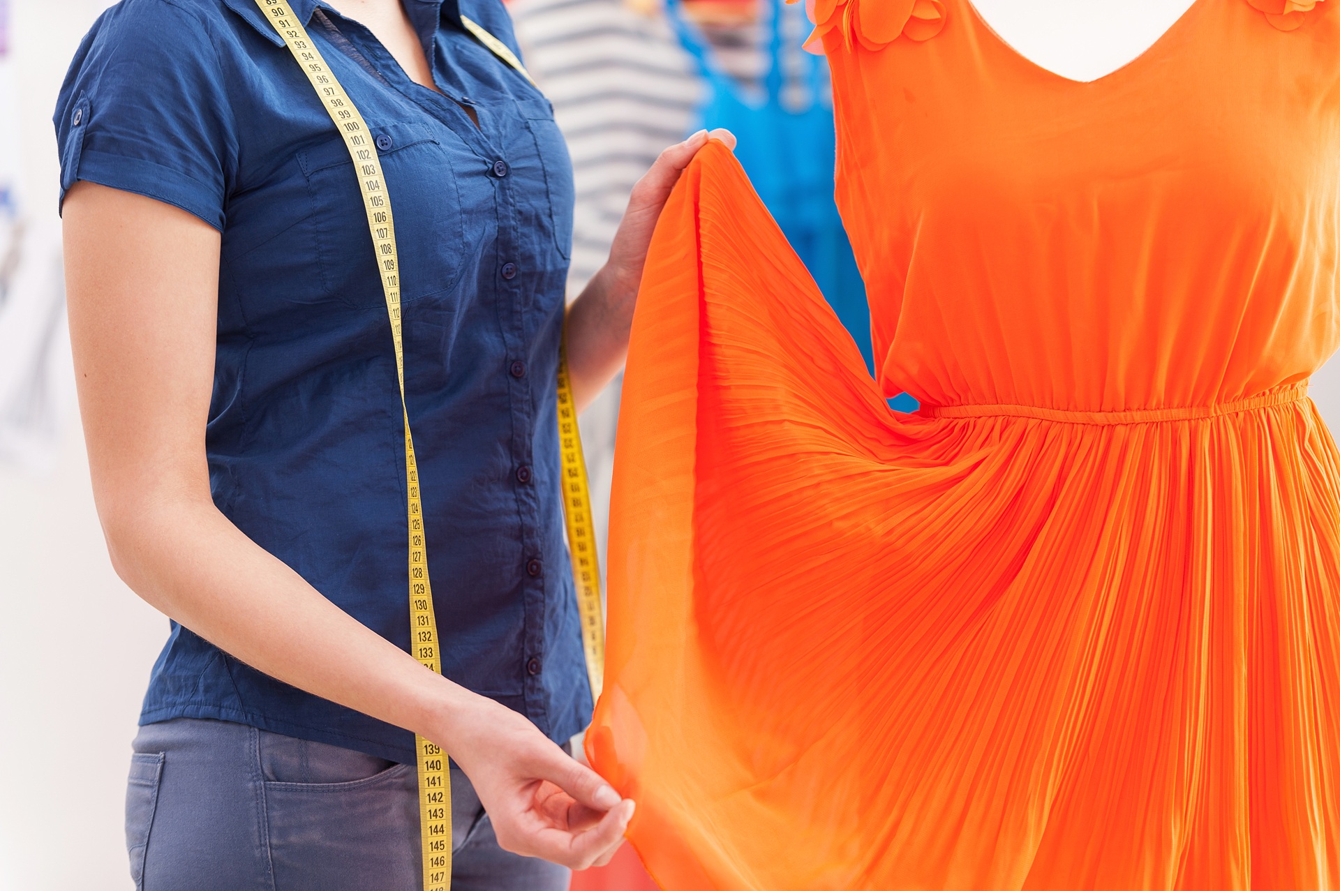 A person holding the skirt of a bright orange dress on a mannequin, with a measuring tape draped around their neck.